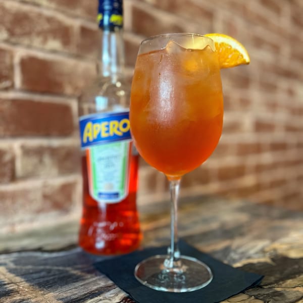 An Aperol Spritz cocktail with an orange slice, next to an Aperol bottle against a brick wall.